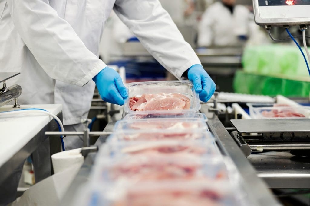 A meat packaging worker handling a small package of chicken tenders on a conveyor belt.