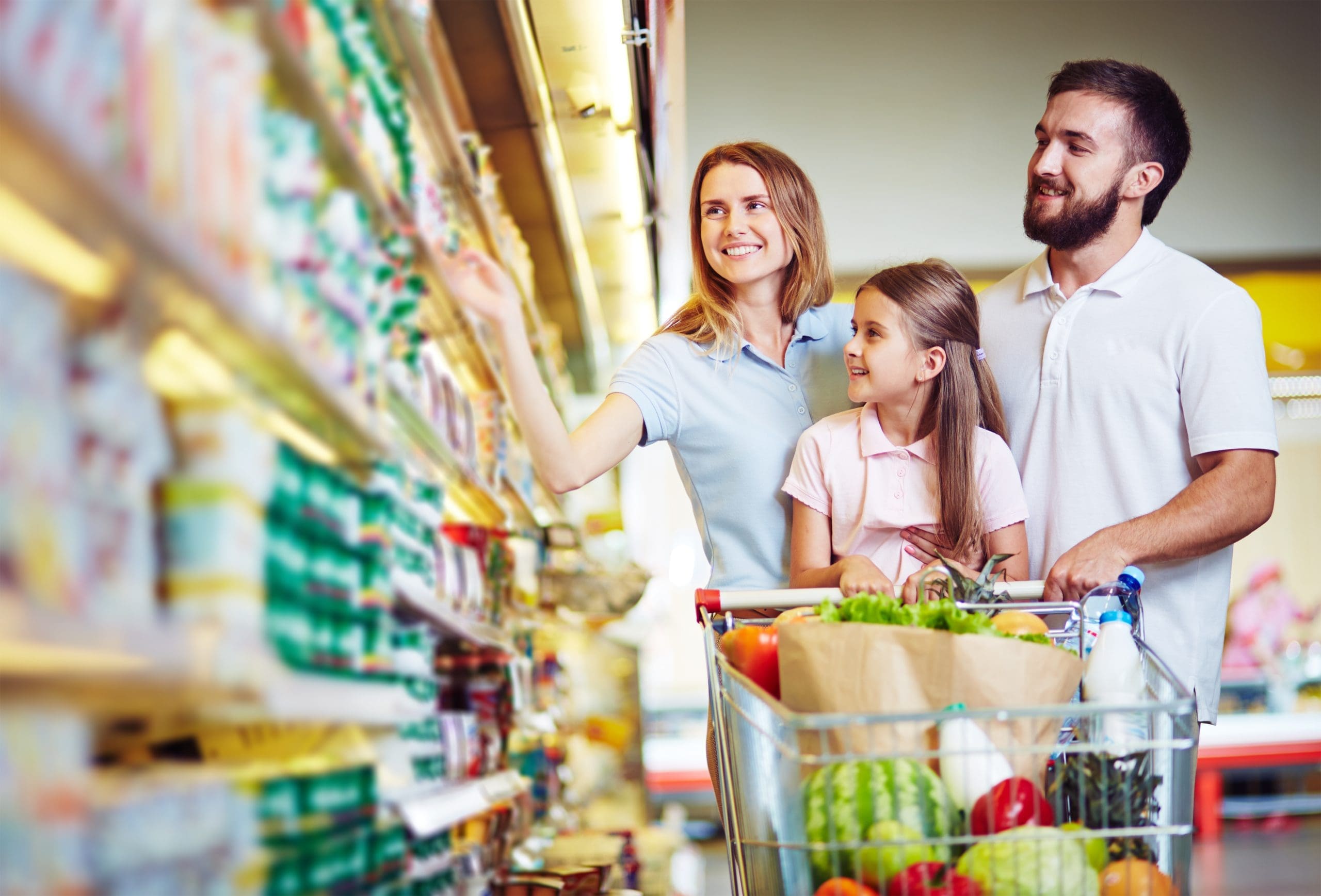 A couple pushing a child in a cart, smiling at a wall of packaged food products in a grocery store.